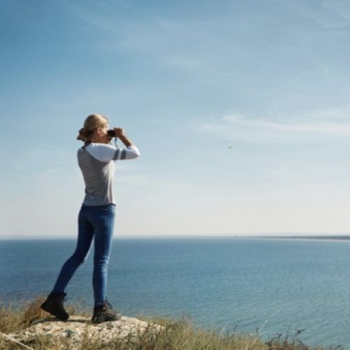 Frau steht auf einer Klippe und schaut durch Fernglas auf's Meer