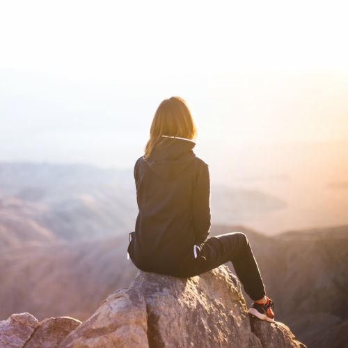 Mädchen sitzt auf Bergspitze, schaut in Richtung Sonnenuntergang